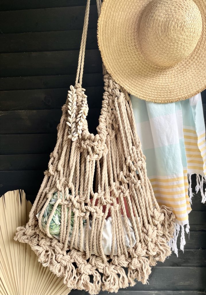 Macrame beach bag hanging in a beach hut with a towel and a palm leaf